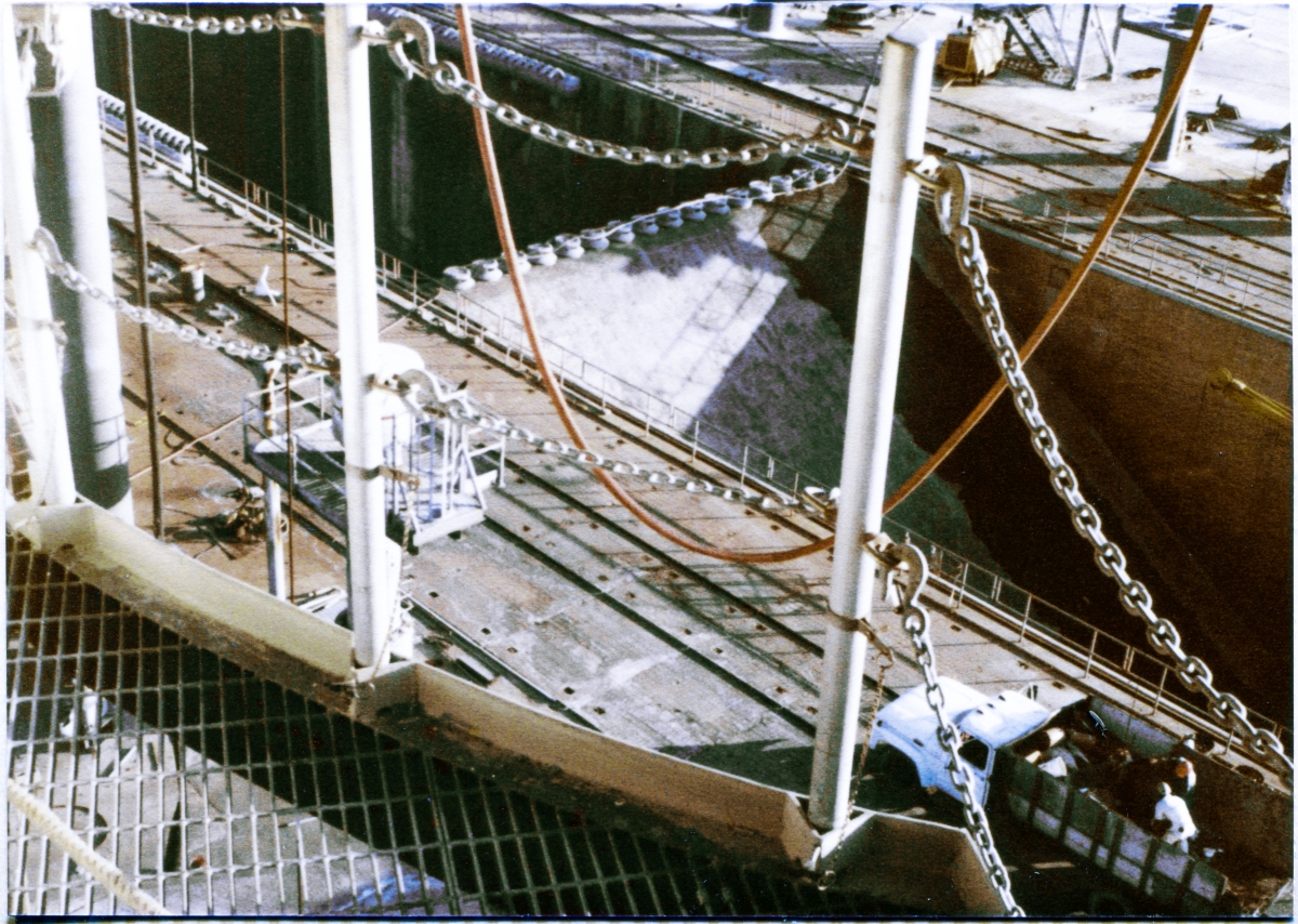 Image 035. The yawning gash of the Flame Trench at Space Shuttle Launch Complex 39-B, Kennedy Space Center, Florida, is filled with the ramping surface of the south side of the Flame Deflector, with its row of Sound Suppression Water Spray Headers along its crest. In this viewpoint, you are standing on the Steel-bar Grating at elevation 112’-0” of the Rotating Service Structure, eye-level over sixty feet above the Pad Deck, looking across and down through Removable Handrail Posts and Safety Chains that guard the curving perimeter of the floor steel in the area of the Orbiter Mold Line for the Space Shuttle’s Right OMS Pod. Photo by James MacLaren.